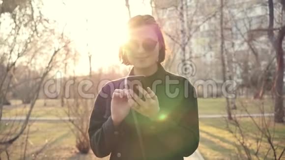 戴着太阳镜微笑的白种人年轻女子在她的智能手机上写了一条信息并在一个城市公园散步慢动作视频的预览图