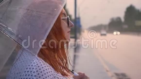 戴眼镜的胖姜女站在公共汽车站看着车撑着伞视频的预览图