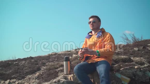 一个穿着黄色夹克蓝色牛仔裤和眼镜的男人坐在山上欣赏风景喝热水瓶里的茶视频的预览图