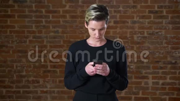 漂亮的白种人金发女性正在滚动她的智能手机同时站在砖背景室内随意的外观视频的预览图