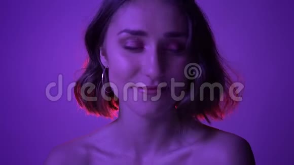 年轻迷人的女性在镜头前用霓虹灯紫色背景微笑的特写镜头视频的预览图
