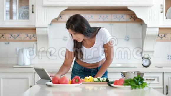 年轻的微笑的女人在厨房里用平板电脑准备食物寻找食谱视频的预览图