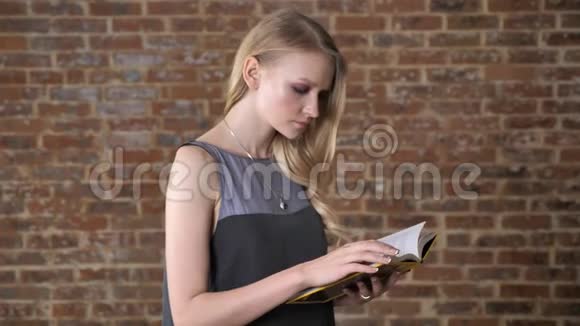 年轻迷人的金发女孩正在看书在镜头前看背景是砖视频的预览图