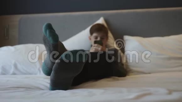 这个男孩躺在床上手里拿着一部智能手机智能手机上的交流和游戏视频的预览图