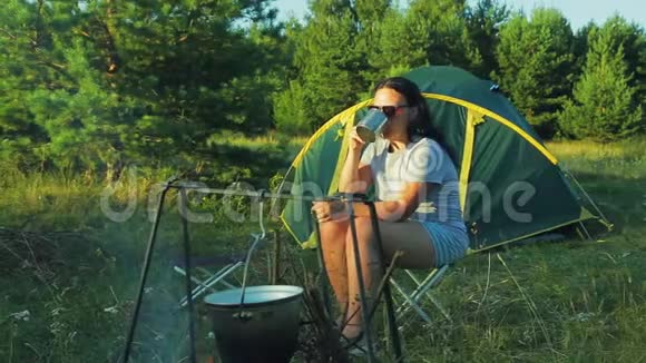 一个戴墨镜的女人坐在火堆旁喝茶欣赏大自然视频的预览图