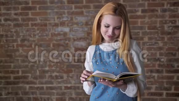 年轻感兴趣的金发女孩正在看书看着摄像机微笑着带着砖头背景视频的预览图