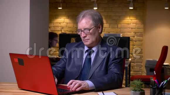 一幅穿着正式服装手提电脑工作的老人在办公室里专心致志疲惫不堪的肖像视频的预览图