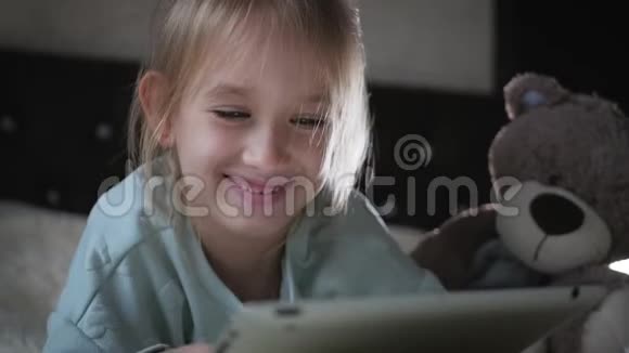 可爱的小女孩坐在床上和泰迪熊一起在平板电脑上玩游戏视频的预览图