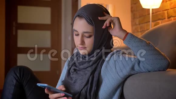 一位年轻迷人的穆斯林女性在坐在地板上的时候在手机上发着头巾的短信视频的预览图