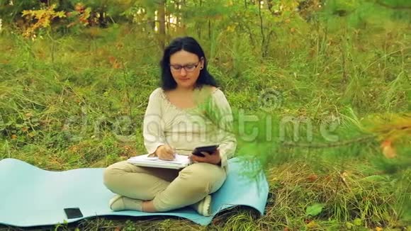 一位戴眼镜的年轻女子在森林边缘的平板电脑上工作并在笔记本上画了一支铅笔视频的预览图