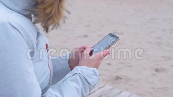 特写女人她的手在社交媒体上在海上背景的电话上打字海上冬天穿白色夹克的女人视频的预览图