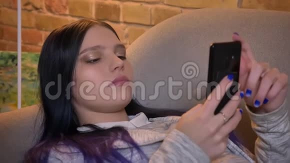 在舒适的家中躺在沙发上用手机发短信的年轻美丽的白种人时髦女性特写镜头视频的预览图