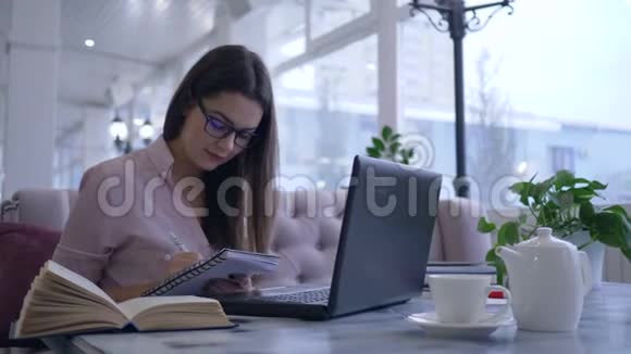 在网上学习女学生在笔记本电脑桌上看书和写笔记视频的预览图