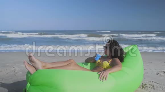 阳光下美丽的女人躺在沙滩上的绿色充气枕头上喝鸡尾酒视频的预览图