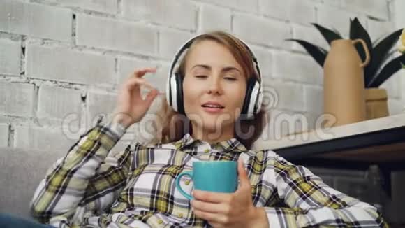 美丽的年轻女子正在唱歌通过耳机听最喜欢的歌拿着一杯热茶现代视频的预览图
