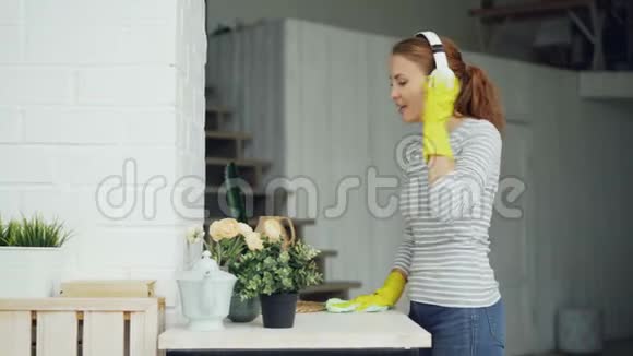 戴着橡胶手套的快乐年轻家庭主妇在家里打扫卫生时通过耳机听音乐唱歌视频的预览图