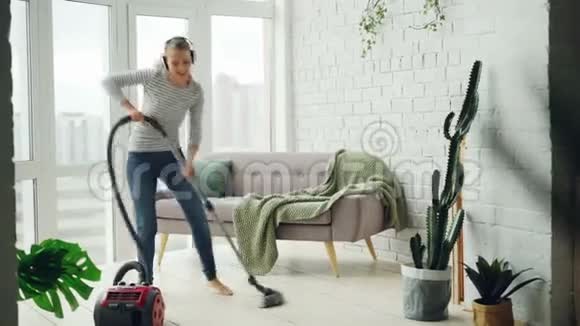 有魅力的年轻女士在家中打扫房间时正在通过耳机跳舞和唱歌女孩是视频的预览图