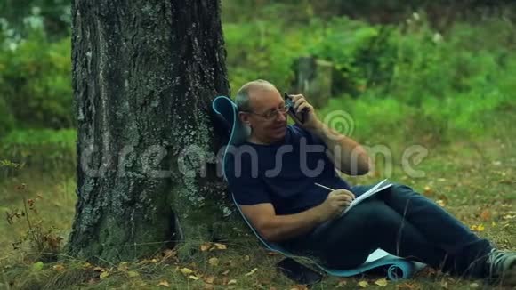 一个戴着眼镜的微笑男子坐在公园的一棵树下用铅笔画一个记事本然后用电话交谈视频的预览图
