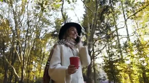 微笑的金发女孩一边喝着咖啡一边在秋天公园的背景上用智能手机交谈视频的预览图