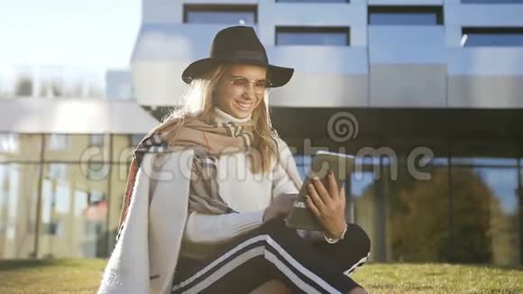 漂亮的女孩戴着时尚的帽子和平板电脑里的人聊天在大楼上的绿草上放松视频的预览图