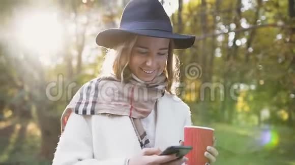 近距离拍摄了一个英俊的女孩戴着帽子和围巾一边喝咖啡一边在公园里散步一边用智能手机视频的预览图