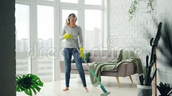 漂亮的女孩正在用扁平的塑料拖把洗地板在耳机里听音乐跳舞和唱歌视频的预览图