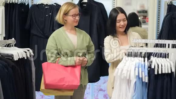 两个年轻的女人穿过商场女人购物年轻女子在服装店挑选服装并咨询视频的预览图