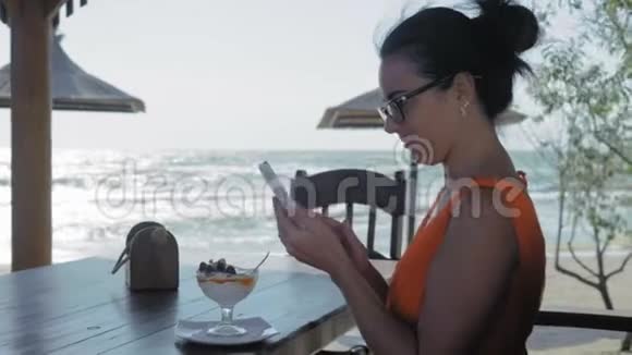 一位戴眼镜的美女坐在海边的咖啡馆里看着智能手机上的照片视频的预览图