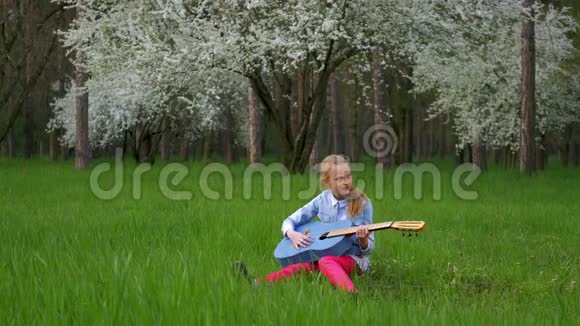 年轻漂亮的金发女孩坐在一个美妙的地方弹吉他在一棵盛开的绿树的背景下视频的预览图