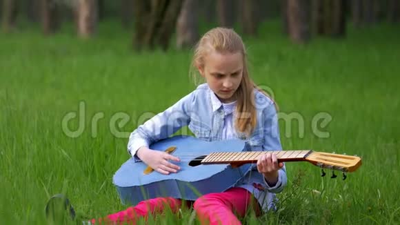 美丽的金发小女孩坐在草地上玩牛仔裤吉他户外乡村的春天视频的预览图