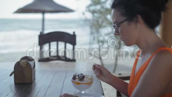 特写镜头戴眼镜的美女坐在海边的木桌旁的咖啡馆里吃甜点视频的预览图