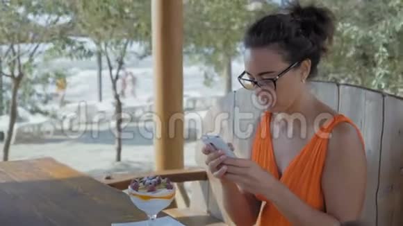 戴眼镜的美女坐在海边的咖啡馆里在智能手机上留言视频的预览图