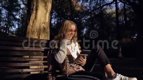 坐在公园的长凳上戴着耳机的年轻英俊的女人用她的平板电脑视频的预览图