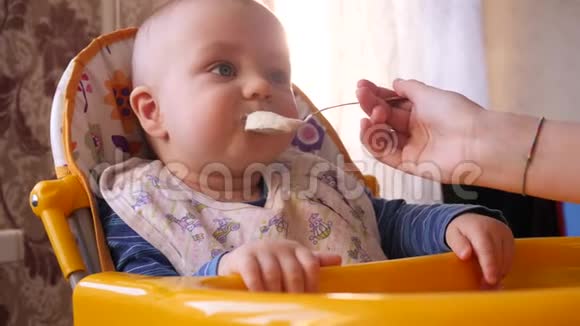 妈妈用勺子在高高的黄色椅子上喂婴儿而小可爱的男孩回头看然后在里面看视频的预览图
