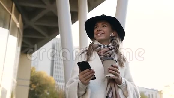 戴着围巾的可爱女孩带着耳机端着一杯咖啡用智能手机聊天视频的预览图
