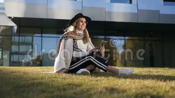年轻的微笑女孩在现代建筑附近的绿草上放松同时使用她的平板电脑视频的预览图