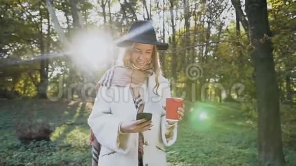 穿着白色外套和帽子的漂亮时尚女人带着咖啡和智能手机在公园里散步视频的预览图