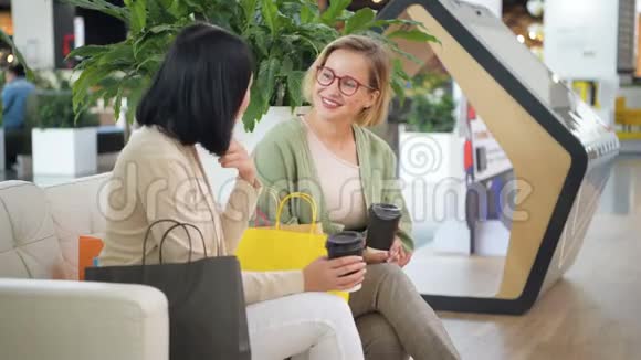 两个年轻的女人在购物后在商场的咖啡馆里喝咖啡谈笑风生人们友谊视频的预览图