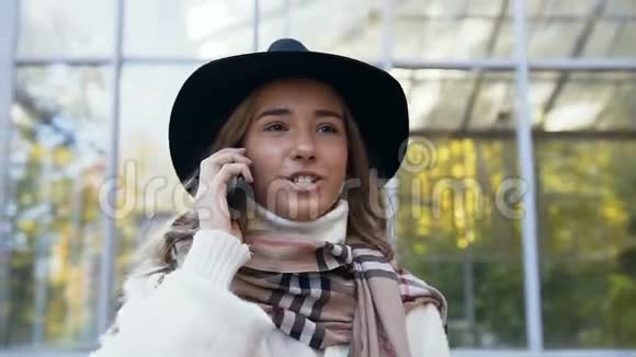 微笑着穿着白色外套的年轻女人在公园里散步用智能手机说话视频的预览图