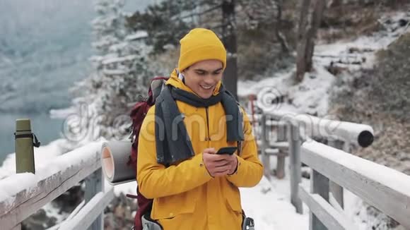 一位年轻的游客站在雪山上的一座桥上使用智能手机背景的美丽山脉视频的预览图