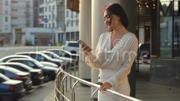 快乐的年轻女子穿着白色衬衫用手机在马路附近的街道上微笑在欧洲城市的汽车视频的预览图