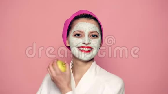 女孩脸上戴着面具头上戴着毛巾手里拿着一个苹果对着粉红色的背景微笑视频的预览图