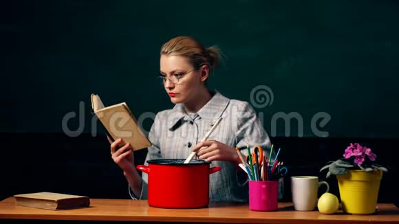 戴眼镜的女人读了一本书在绿色学生板的背景下用红锅里的勺子混合了一些东西视频的预览图