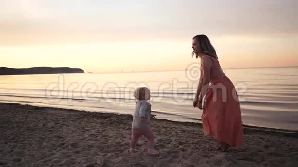 日落时分穿着裙子的年轻妈妈和一个小女儿沿着海滨散步视频的预览图