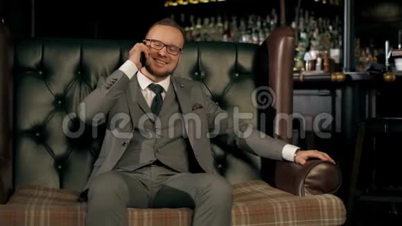 成功的胡子商人穿着西装坐在豪华的皮革沙发上打电话享受成功视频的预览图