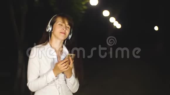 戴着耳机听歌唱歌的女人通过触摸电话在城市公园里唱歌视频的预览图