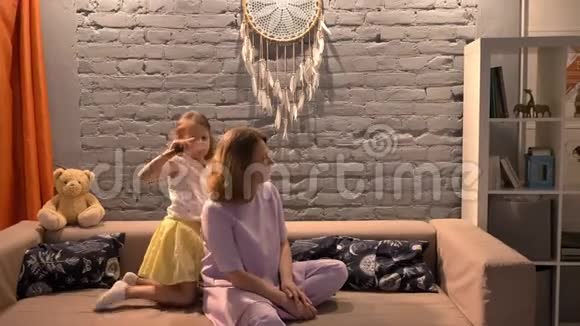小女儿用梳子梳母亲的头发坐在现代客厅的沙发上家庭观念在室内视频的预览图