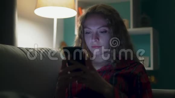 美丽的年轻女性晚上在舒适的房间里的沙发上使用智能手机视频的预览图
