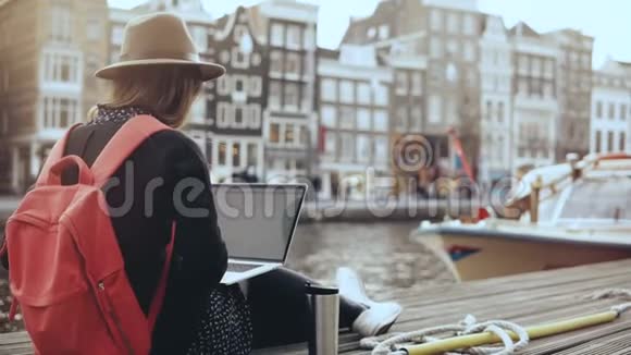 4K30的快乐女商人用笔记本电脑工作阿姆斯特丹带红色背包的女士坐在美丽的河堤上视频的预览图