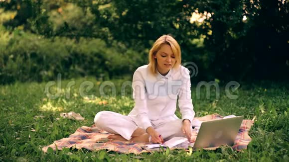 女孩在公园里用笔记本电脑坐在毯子上带着笔记本电脑在户外学习的年轻女人视频的预览图